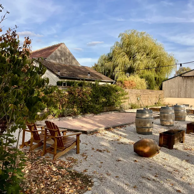 Le terrain de pétanque en gravier, ses fauteuils en bois et ses tonneaux, devant le saule pleureur