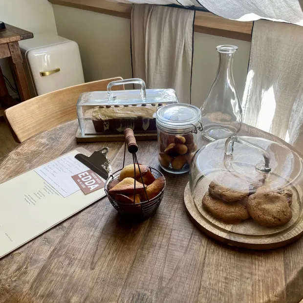 Une table ronde en bois dressée pour le petit-déjeuner, cake sous cloche et bocal de biscuits
