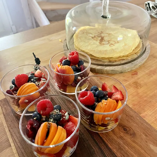 Trois coupes de salade de fruits frais et une pile de crêpes sous cloche, sur une table de bois clair
